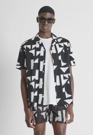 Jonge man met een zwart-wit geometrisch patroon kortemouwen shirt en bijpassende korte broek, met een wit T-shirt en zwarte zonnebril.