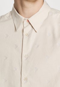 Primer plano de una camisa beige con botones, cuello clásico y sutiles patrones tejidos en forma de pájaros en la tela.