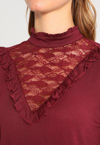Burgundy-paita, jossa on korkea röyhelökaulus, pitsipaneeli ja pitsiylitys kukkakuviolla, asettuneena sileän kankaan päälle.