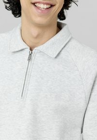 Personne souriante portant un pullover léger gris clair à col zippé sur un fond uni.