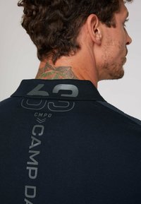 Granatowa koszulka polo z teksturowanej tkaniny, z prążkowanym kołnierzykiem, dużym szarym nadrukiem oraz detalami brandingowymi z tyłu.