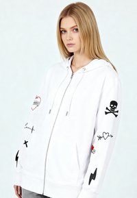 Felpa con cappuccio bianca dotata di cerniera integrale, cappuccio e molteplici motivi grafici neri e rossi, tra cui cuori, un teschio e fulmini.