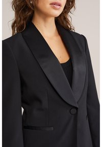 Kvinna som bär en skräddarsydd svart blazer med satinlappar och en täckt knapp över en svart topp, visas från nacke till midja.