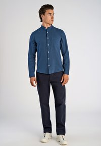 Camicetta a maniche lunghe, blu scuro, con bottoni e una piccola tasca, indossata con pantaloni scuri e sneakers bianche, che mostrano una texture liscia.