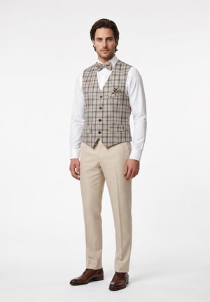 Mann trägt beige karierte Weste und Fliege, weißes Hemd, beige Hose und braune Anzugsschuhe, steht vor einem einfarbigen weißen Hintergrund.