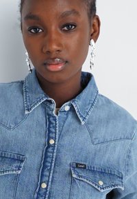 Chemise en denim bleu au design classique, avec un col pointu, des poches avant et une fermeture à boutons. Des boucles d'oreilles argentées ajoutent du contraste.