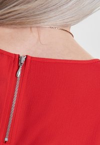 Vêtement rouge en tissu texturé, avec une fermeture éclair argentée visible au niveau de l'encolure arrière, col rond à finition lisse.