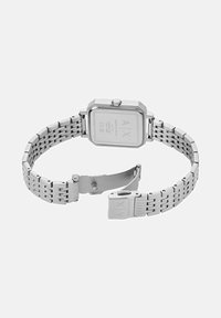 Reloj de pulsera con brazalete de metal plateado, caja cuadrada y un cierre abierto que muestra la parte inferior grabada con texto de marca.