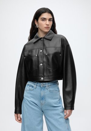 Ung kvinde iført en sort tekstureret cropped jakke og lyseblå højtaljede jeans, stående mod en ensfarvet lys baggrund.