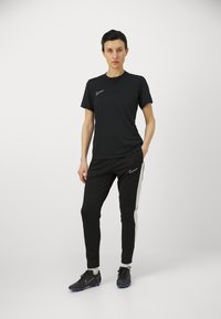 Crna sportska majica i hlače imaju mali Nike logo. Hlače imaju bijelu bočnu prugu i uparene su s crnim sportskim cipelama.