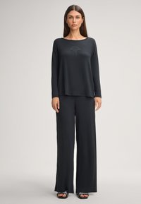 Czarna bluzka z długim rękawem, o zaokrąglonym dekolcie i subtelnym dekoracyjnym wzorze, zestawiona z czarnymi spodniami o szerokich nogawkach i płaskimi sandałami.