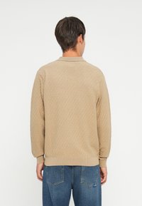 Beżowy sweter w wzory z długimi rękawami i ściągaczami, z klasycznym kołnierzykiem i luźnym krojem, w połączeniu z niebieskimi dżinsami.