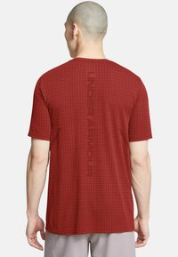 Czerwona koszulka z krótkim rękawem o teksturowanym wzorze siatki, z napisem "UNDER ARMOUR" drukowanym pionowo w ciemnoczerwonym kolorze na plecach.