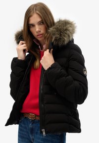 Zwarte pufferwinterjas met een faux fur gevoerde capuchon, ritssluiting en zijzakken. Draagt over een rode trui en een blauwe spijkerbroek.