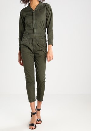 Kobieta w oliwkowozielonym kombinezonie z długim rękawem, z paskiem w talii i czarnych sandałkach na wysokim obcasie z paskiem wokół kostki na jednolitym tle.