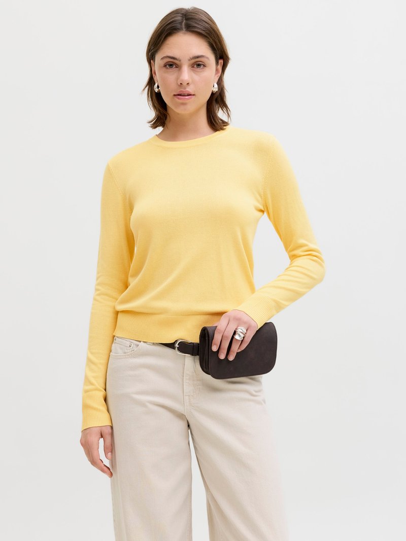 Vrouw draagt een geel lange-mouwen trui en beige broek, houdt een kleine zwarte leren clutch tasje bij haar taille vast.
