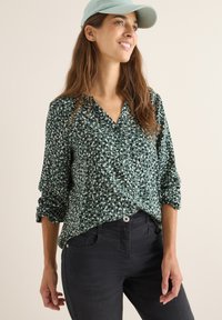Grün gemusterte Bluse mit V-Ausschnitt und Knopfdetail, kombiniert mit dunklen Jeans. Der Stoff hat ein leichtes, leicht strukturiertes Aussehen.
