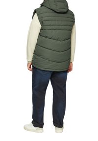 Grüne gepolsterte Weste mit Kapuze, mit horizontalen Nähten. Kombiniert mit einem cremefarbenen Langarmshirt und dunklen Jeans. Weiße Sneaker sind sichtbar.