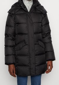 Zwarte pufferjas met een capuchon, uitgerust met drukknoopsluiting, opgestikte zakken en een gewatteerd ontwerp voor warmte en isolatie. Zachte textuur.