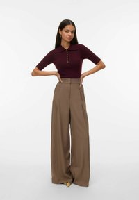 Mujer con cabello largo y oscuro que lleva una blusa corta burdeos y pantalones marrones de tiro alto y pierna ancha, de pie con las manos en las caderas.