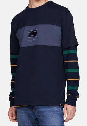 Chemise à manches longues marine avec un design en color-block, panneaux bleu foncé et gris, manches rayées vertes et orange, et logo sur la poitrine.