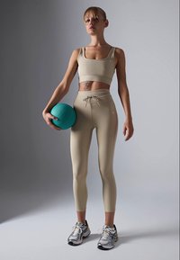 Kvinne i beige sportsbekledning som holder en tealfarget medisinball, står med en selvsikker holdning mot en ensfarget grå bakgrunn.