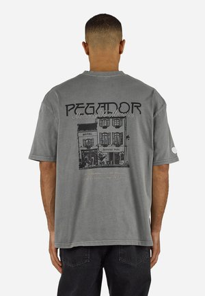 Muž otočený chrbtom, ktorý nosí sivé nadrozmerné tričko s čiernym náčrtom budovy a textom "PEGADOR Moments To Memories" na zadnej strane.