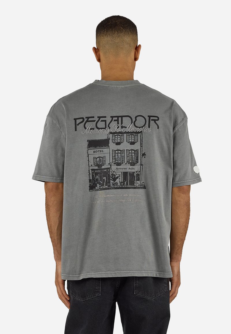 Άνδρας με γκρι υπερμεγέθη μπλουζάκι που απεικονίζει ένα σκίτσο μαύρου κτιρίου και κείμενο στο πίσω μέρος που γράφει «PEGADOR Moments To Memories».