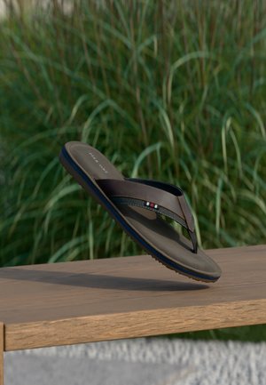 Καφέ και μαύρη σαγιονάρα με μπλε λεπτομέρεια στη σόλα, κρεμασμένη πάνω από ξύλινο παγκάκι με φόντο πράσινο γρασίδι.