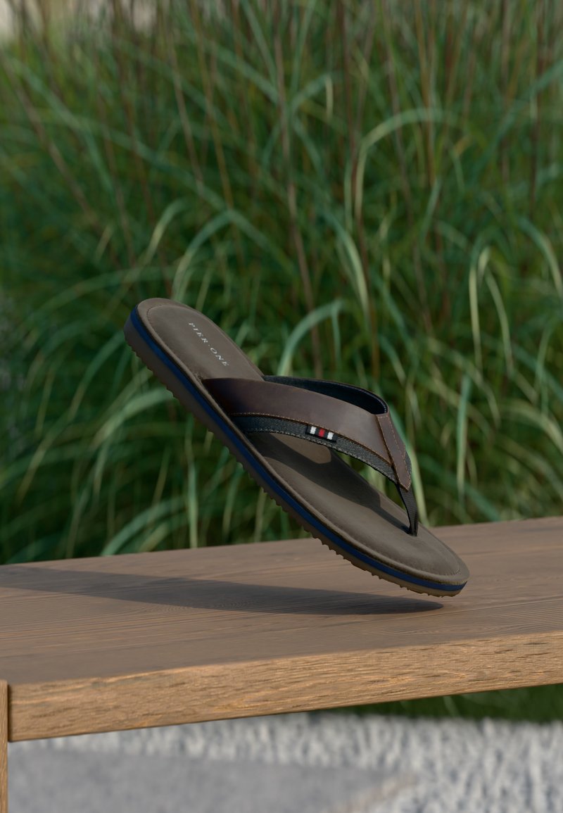 Καφέ και μαύρη σαγιονάρα με μπλε λεπτομέρεια στη σόλα, κρεμασμένη πάνω από ξύλινο παγκάκι με φόντο πράσινο γρασίδι.