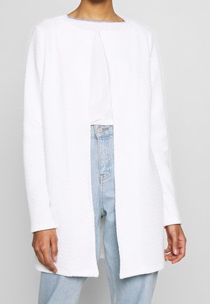 Personne portant une veste blanche texturée à manches longues ouverte sur le devant, sur un top blanc et un jean taille haute bleu clair.