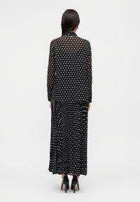 Μαύρο διάφανο πουκάμισο με λευκές πουά, μακριά μανίκια, μανσέτες με κουμπιά, σε συνδυασμό με μαύρη μακριά φούστα με πουά και πιέτες.