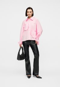 Γυναίκα που φοράει ροζ μπλούζα με διάφορα βολάν σε σχήμα καρδιάς, μαύρο δερμάτινο παντελόνι, μαύρα μυτερά τακούνια και κρατάει μια μαύρη τσάντα με καρφιά.