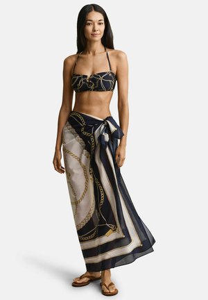 Femme portant un haut de bikini noir à imprimé chaîne dorée avec un grand paréo assorti noué à la taille et des sandales tongs marron.