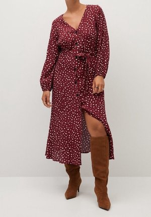Vrouw draagt een bordeauxrode, lange mouwen, geknoopte jurk met witte stippen, een tailleceintuur en bruine suède laarzen tot over de knie.