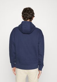 Marinblå munkjacka med huva, tillverkad av mjuk bomull, med en avslappnad passform, ribbade ärmslut och midja för en snug avslutning.