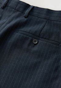 Pantalon bleu marine foncé à fines rayures, avec une poche passepoilée arrière boutonnée et une boucle de ceinture sur fond blanc.