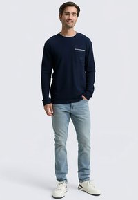 Navy long-sleeve t-shirt with a front pocket, paired with light blue jeans and white sneakers. Smooth fabric, crew neck design, simple accents.