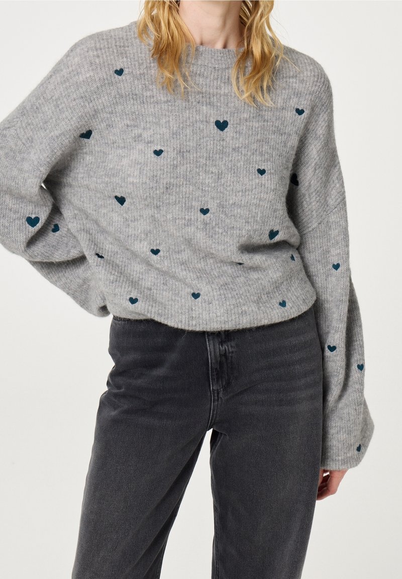 Femme portant un pull en maille gris avec de petits motifs en forme de cœur foncés et un jean noir taille haute, posant avec une main sur la hanche.