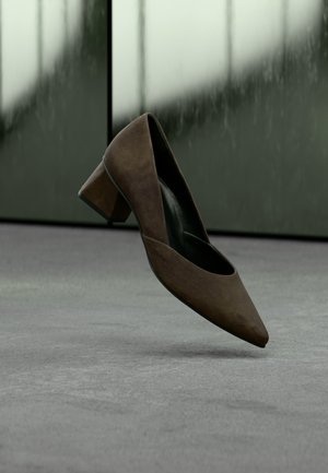 Brauner Damensandalette aus Wildleder mit Blockabsatz, schwebend über einer grauen Fläche, unscharfer dunkelgrüner und grauer Hintergrund.