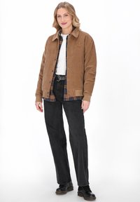 Kvinde står iført en tan corduroyjakke over en ternet skjorte, hvid top, sorte bukser, sort bælte og sorte støvler på en hvid baggrund.