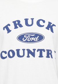 Hvit bomull t-skjorte med store blå bokstaver som leser "TRUCK COUNTRY" over en blå Ford-logo, med et slitt utseende.
