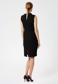 Vestido negro sin mangas, ajustado, con cuello, detalle de abertura en la espalda, tejido suave, hasta los tobillos, combinado con tacones altos negros.