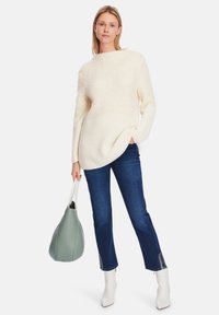 Cream fuzzy knit sweater with ribbed hem, paired with dark blue jeans featuring lace-up accents at the cuffs. Holding a soft green handbag.