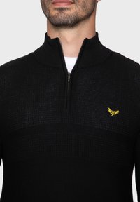 Schwarzer Strickpullover mit einem halben Reißverschluss am Kragen, der ein strukturiertes Muster aufweist und ein kleines gelbes Logo auf der linken Brust hat.
