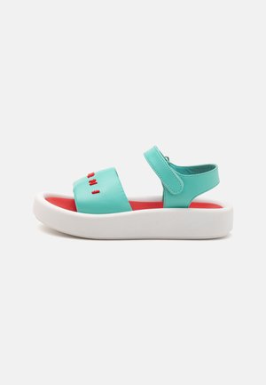Turquoise sandaaltjes met een witte zool, voorzien van een brede band en een klittenbandsluiting. Rode tekstaccenten op de bovenste band. Soepele textuur.