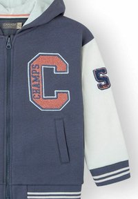 Chaqueta con capucha de cremallera en azul marino y blanco, con bordados de los acentos "C" y "S", bolsillos laterales y puños y dobladillo a rayas.