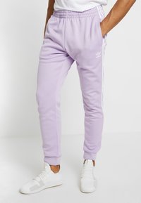 Lila Adidas-joggingbyxor med elastiskt midjeband, sidofickor, vita ränder längs sidorna och muddar vid anklarna, matchade med vita sneakers.