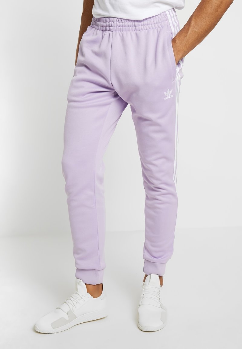 Lila Adidas-joggingbyxor med elastiskt midjeband, sidofickor, vita ränder längs sidorna och muddar vid anklarna, matchade med vita sneakers.