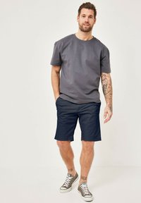Graues Baumwoll-T-Shirt mit kurzen Ärmeln, kombiniert mit marineblauen Shorts. Das Modell trägt graue Sneaker und zeigt einen lässigen, entspannten Look.